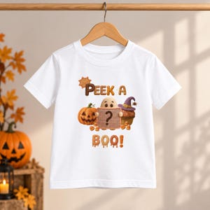 Könnte beinhalten: Ein weißes Kurzarm-T-Shirt mit einem Grafikdruck eines Geistes, der ein Schild mit einem Fragezeichen hält, einem orangefarbenen Kürbis und einem Cupcake mit einem violetten Hexenhut. Der Text lautet PEEK-A-BOO in braunen und orangen Buchstaben.