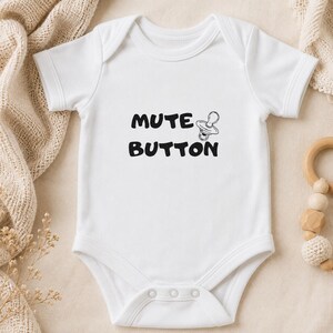 May include: A white short-sleeved baby bodysuit featuring the black text MUTE BUTTON and a graphic illustration of a pacifier. The garment has a round neckline and three snap closures at the bottom for easy access, designed for infants.