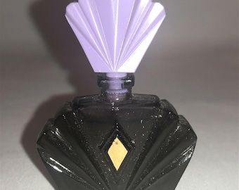 May include: A black glass perfume bottle with a faceted, fan-shaped body and a central diamond-shaped gold inlay. The bottle features a light purple, fan-shaped stopper that sits atop the narrow neck, creating a distinct geometric silhouette.