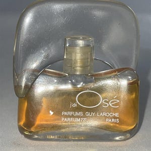 May include: A clear glass perfume bottle with a unique curved top piece and amber-colored liquid inside. The front features white text reading j'ai Osé PARFUMS GUY LAROCHE PARFUM 77 PARIS. The bottle has a small, transparent cylindrical cap on top.