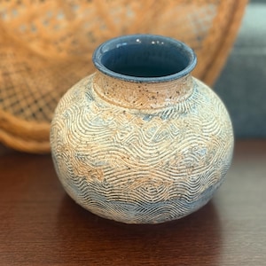 May include: A round ceramic vase with a narrow neck and a wide body. The exterior features a textured, wavy line pattern in shades of tan and blue. The rim is finished in a smooth, solid blue glaze, while the body displays a speckled, earthy finish.