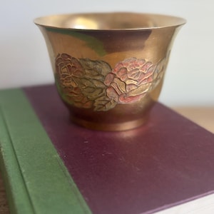 Könnte beinhalten: Eine Messingschale mit ausgestelltem Rand und konischem Fuß. Die Außenseite ist mit einem geprägten Blumenmuster versehen, das handbemalte Details in gedeckten Rosa-, Grün- und Brauntönen aufweist und stilisierte Blumen und Blätter zeigt.