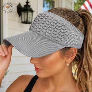 Puede incluir: Una visera de punto gris con un patrón de trenzas texturizado en el panel frontal, una visera acanalada y una estructura de punto tipo malla en los paneles laterales. La visera incluye un parche circular con el texto HATHATCH HANDCRAFTED HEADWEAR.