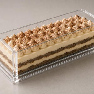 Op de afbeelding: Een rechthoekige, doorzichtige acryl bak met een gelaagd tiramisu dessert. Het dessert bestaat uit afwisselende lagen in koffie gedrenkte cake en room, afgewerkt met toefjes room bestrooid met een fijn laagje bruin cacaopoeder.