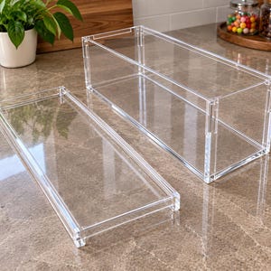 Könnte beinhalten: Eine rechteckige Aufbewahrungsbox aus klarem Acryl mit einem passenden abnehmbaren Deckel. Der Behälter zeichnet sich durch ein transparentes Design mit sichtbaren ineinandergreifenden Verbindungen an den Ecken aus, die einen klaren Blick ins Innere ermöglichen.
