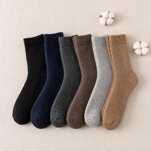 May include: A set of six crew socks in a row, featuring a ribbed cuff and a soft, textured knit finish. The colours include black, navy blue, charcoal grey, dark brown, light grey, and tan, accompanied by two small white cotton bolls.