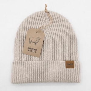 May include: A beige ribbed knit beanie made of merino wool. A brown paper tag attached with twine features a line drawing of a sheep and the text MERINO WOOL. A small brown rectangular patch on the folded cuff displays the brand name SockE.