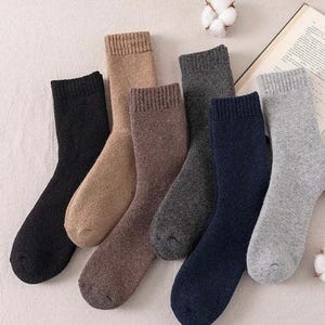 May include: A set of six crew-length socks in solid colours including black, tan, brown, charcoal grey, navy blue, and light grey. Each sock features a ribbed cuff at the top and a soft, textured knit construction, arranged in a fan-like pattern.