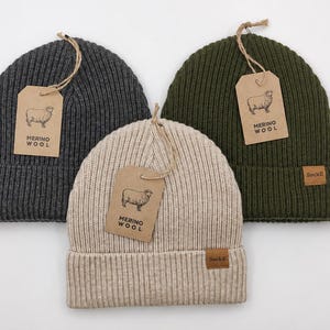 Puede incluir: Tres gorros de punto acanalado en gris carbón, verde oliva y beige. Cada gorro tiene un dobladillo con una pequeña etiqueta rectangular marrón que dice SockIt y una etiqueta de papel marrón con un dibujo de una oveja y MERINO WOOL.