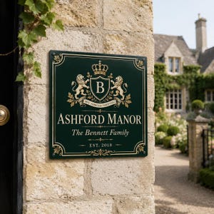 Puede incluir: Un letrero cuadrado de color verde oscuro con letras doradas y bordes decorativos. El letrero presenta un escudo central con una corona, dos leones y la letra B, con el texto Ashford Manor, The Bennett Family y Est. 2018 impreso en oro.
