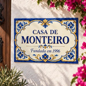 Könnte beinhalten: Eine rechteckige Wandplakette aus Keramik mit cremefarbener Mitte und einem dekorativen blau-goldenen Rand. Der Text lautet CASA DE MONTEIRO und Fundada em 1996 in dunkelblauer Schrift, verziert mit floralen Ornamenten in den Ecken.