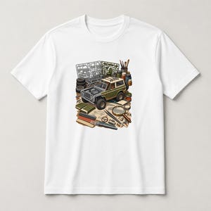 May include: A white short-sleeve t-shirt featuring a graphic illustration of an olive green off-road vehicle model kit. The design includes plastic parts on sprues, paint bottles, a magnifying glass, and various modeling tools arranged on a workbench.