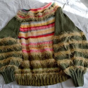 Könnte beinhalten: Ein langärmeliger Strickpullover mit olivgrünen Ärmeln und einem Mittelteil mit horizontalen Streifen in Pink, Gelb und Weiß. Die Ärmel und der untere Teil sind mit horizontalen Reihen aus beigefarbenen, flauschigen Garnfransen verziert.
