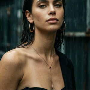 May include: A pair of gold-toned hoop earrings and a matching gold-toned lariat necklace featuring a delicate chain, a small spherical bead, and a slender, elongated drop pendant. The jewellery set is worn against a dark background with rain streaks.