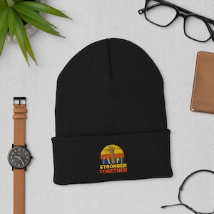 May include: A black cuffed beanie featuring an embroidered graphic of a yellow sun with orange and red stripes behind several raised hands. Below the graphic, the text reads STRONGER TOGETHER in orange capital letters on the front of the hat.