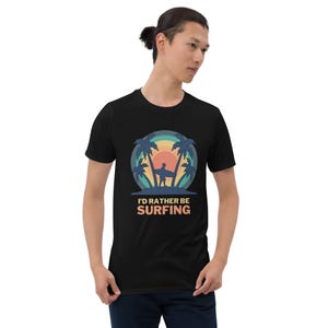 Peut inclure: Un t-shirt noir à manches courtes avec un motif circulaire représentant un surfeur entre deux palmiers devant un coucher de soleil. Sous le motif, le texte I'D RATHER BE SURFING est écrit dans une police grasse avec des tons rétro.