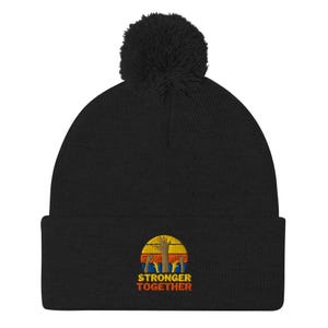 May include: A black knit beanie with a pom-pom on top. The front features an embroidered graphic of a sunset in yellow, orange, and red stripes behind several raised hands. Below the graphic, the text reads STRONGER TOGETHER in orange letters.