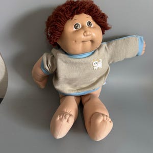 Könnte beinhalten: Eine Puppe mit einem weichen, hellen Stoffkörper, rundem Gesicht und Grübchen an den Wangen. Sie hat kurzes, lockiges braunes Wollhaar und trägt ein beiges Langarmshirt mit hellblauen Abschlüssen und einer kleinen weißen Katzenstickerei.