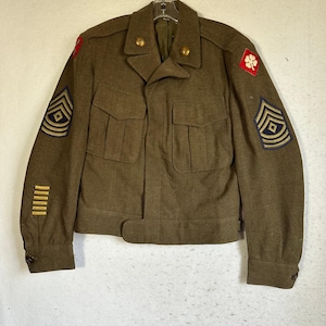 Può includere: Una giacca militare in lana color verde oliva con due tasche sul petto, bottoni dorati e toppe ricamate su entrambe le maniche. La giacca presenta gradi a gallone, strisce di servizio sulla manica inferiore e una toppa a trifoglio rosso.