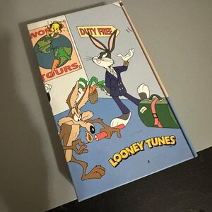 May include: A Looney Tunes VHS tape case featuring Bugs Bunny in a pilot uniform with a green suitcase, Wile E. Coyote holding a lit stick of dynamite, and Tweety Bird on a globe. The text reads World Tours, Duty Free, and Looney Tunes.