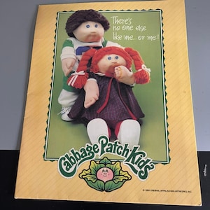 Puede incluir: Un anuncio rectangular de Cabbage Patch Kids con dos muñecos sobre un fondo verde. Uno tiene el pelo castaño oscuro y una camiseta verde y blanca, el otro trenzas rojas, un vestido de cuadros oscuros y un chupete. El texto dice: There's no one else like me... or me!