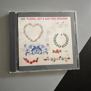 May include: A clear plastic CD case containing a white insert titled 102 FLORAL ART & QUILTING DESIGNS. The insert features various colorful embroidered-style illustrations, including a heart-shaped wreath, blue floral motifs, and green leaves.
