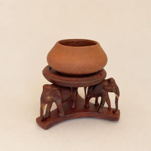 Op de afbeelding: Een kleine, ronde kom in terracottakleur rust op een donkerbruine houten standaard. De standaard is voorzien van drie uitgesneden olifantenfiguren die een cirkelvormig platform ondersteunen, elk geplaatst op een gebogen basissegment.