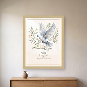 May include: A framed wall art print featuring a watercolor illustration of a white dove surrounded by green leaves and small yellow flowers. The text reads Celebration of Faith, Anna, 12 June 2027, Lisbon, and Always in our hearts in black font.