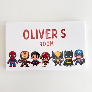 May include: A rectangular white sign featuring the red text OLIVER'S ROOM at the top. Below the text are eight colourful cartoon-style superhero figures including Spider-Man, Iron Man, Superman, The Flash, Wolverine, Batman, and Captain America.