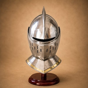 May include: A polished steel knight helmet featuring a central ridge, a horizontal visor slit, and vertical ventilation openings. The base of the helmet is accented with a gold-colored metal trim secured by rivets, mounted on a dark wooden stand.