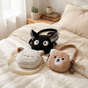 Peut inclure: Trois sacs à bandoulière en peluche avec des visages d'animaux. Un chat noir aux grands yeux blancs, un mouton gris clair aux yeux fermés et oreilles fauves, et un ours fauve avec un museau blanc, un nez noir et une petite langue rose.