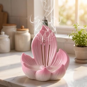 Op de afbeelding: Een roze keramische wierookhouder in de vorm van een lotusbloem met drie dunne wierookstokjes die rechtop in het midden staan. Witte rookpluimen stijgen op van de brandende uiteinden van de stokjes en krullen in de lucht boven de bloemblaadjes.