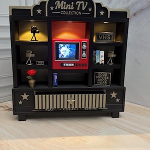 Può includere: Uno scaffale espositivo in legno nero etichettato Mini TV Collection con accenti dorati, stelle e uno schermo televisivo rosso centrale. Gli scaffali contengono oggetti in miniatura a tema cinematografico, tra cui macchine fotografiche, cassette VHS, una rosa rossa e un cartello con la scritta Good Movies Good Mood.
