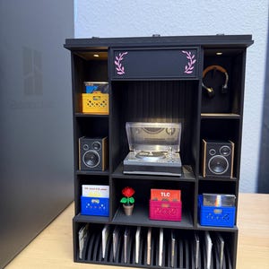 May include: A black miniature shelving unit featuring a central record player, two speakers, and various storage compartments. It contains small crates holding records, a potted rose, headphones, and a bottom row of vertical slots for media cases.