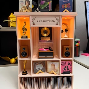 Può includere: Uno scaffale musicale in miniatura rosa chiaro con un giradischi centrale, altoparlanti, trofei dorati e varie copertine di album. Il pannello superiore mostra il testo SURTI EFFECTS 3D. L'unità include un'illuminazione calda integrata.