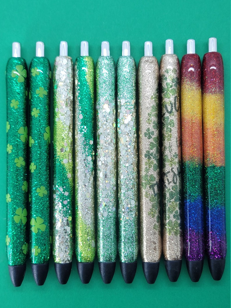 May include: Ten glitter pens with different designs. The pens have a clear plastic body and a black cap. The glitter designs include green shamrocks, green and gold glitter, and a rainbow.