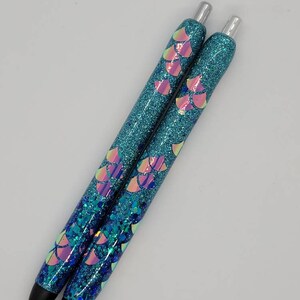 May include: Two blue glitter pens with iridescent mermaid scale designs. The pens have silver caps and black ink.