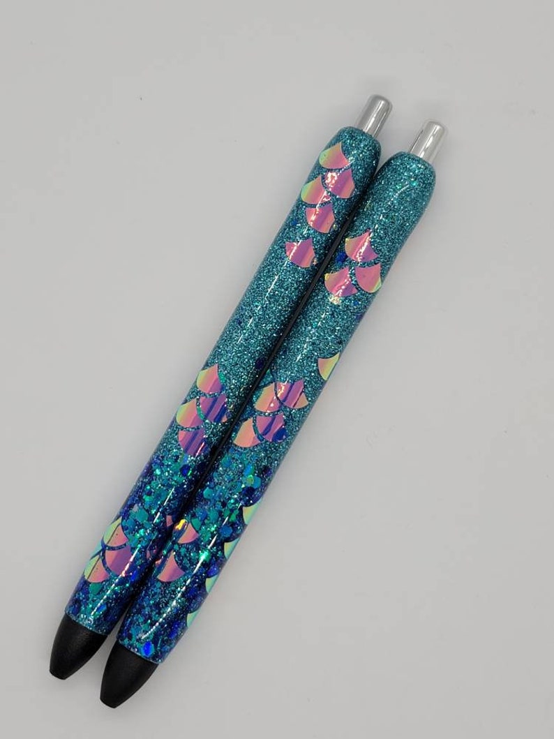 May include: Two blue glitter pens with iridescent mermaid scale designs. The pens have silver caps and black ink.