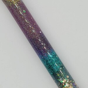 May include: A close-up of a glittery, iridescent pen with a gradient of colors from gold to purple to teal. The pen is made of clear resin and has a variety of glitter embedded in it.