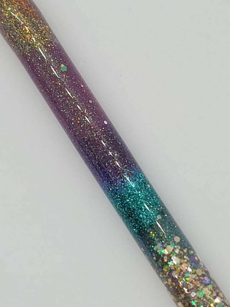May include: A close-up of a glittery, iridescent pen with a gradient of colors from gold to purple to teal. The pen is made of clear resin and has a variety of glitter embedded in it.