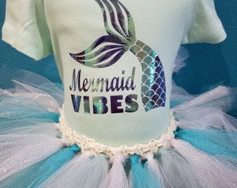 Peut inclure: Un body bébé vert clair avec une queue de sirène holographique et le texte "Mermaid Vibes". Le body est associé à un tutu en tulle blanc et turquoise.