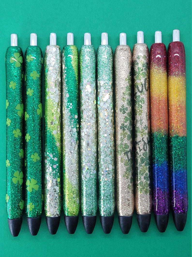 May include: Ten glitter pens with different designs. The pens have black caps and silver accents. The designs include green shamrocks, green and silver glitter, iridescent glitter, gold glitter, and rainbow glitter.