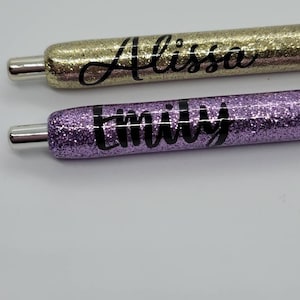 May include: Two glitter pens with names written on them. The top pen is gold glitter with the name "Alissa" written in black. The bottom pen is purple glitter with the name "Emily" written in black.