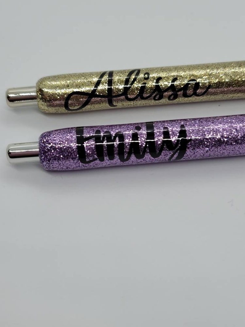 May include: Two glitter pens with names written on them. The top pen is gold glitter with the name "Alissa" written in black. The bottom pen is purple glitter with the name "Emily" written in black.