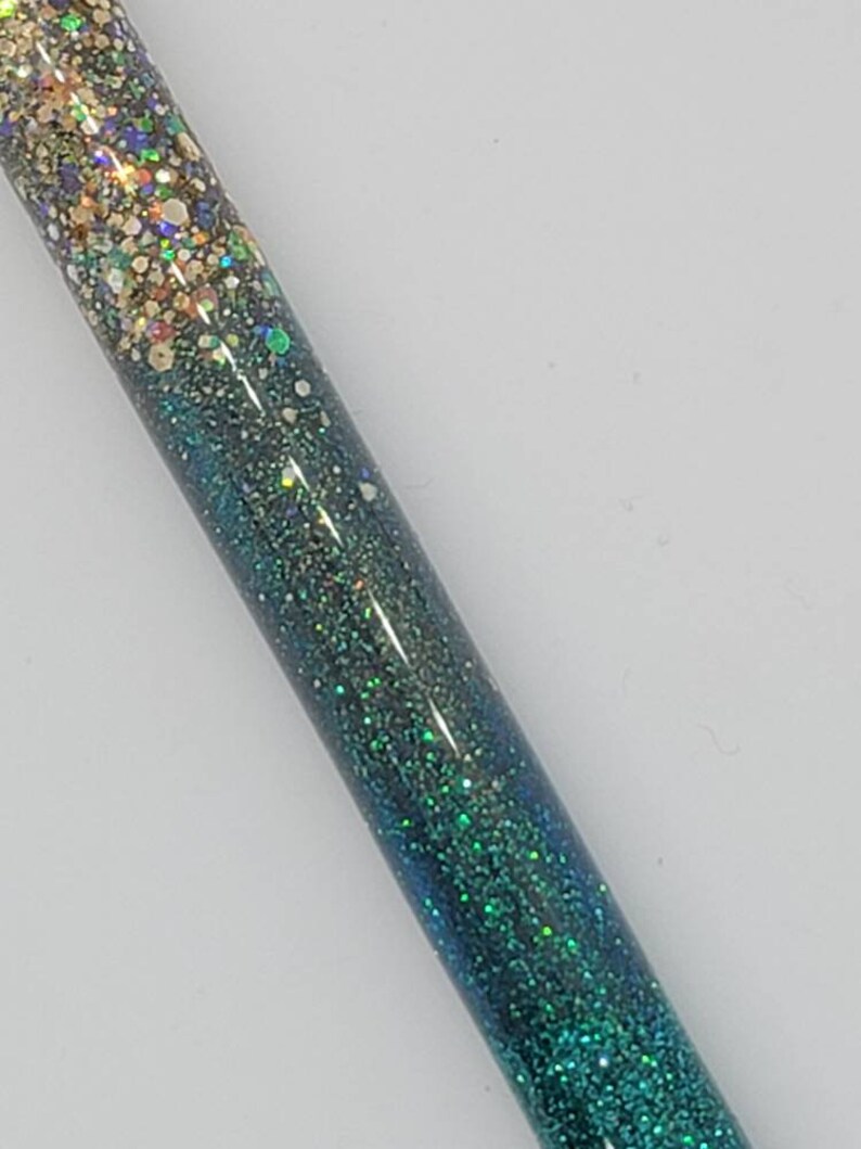 May include: A close-up of a green and gold glitter pen. The pen has a gradient of glitter, starting with gold at the top and fading to green at the bottom.