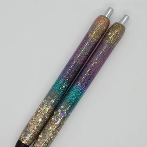 May include: Two iridescent glitter pens with a gradient of purple, teal, and gold glitter. The pens have silver caps and black ink.