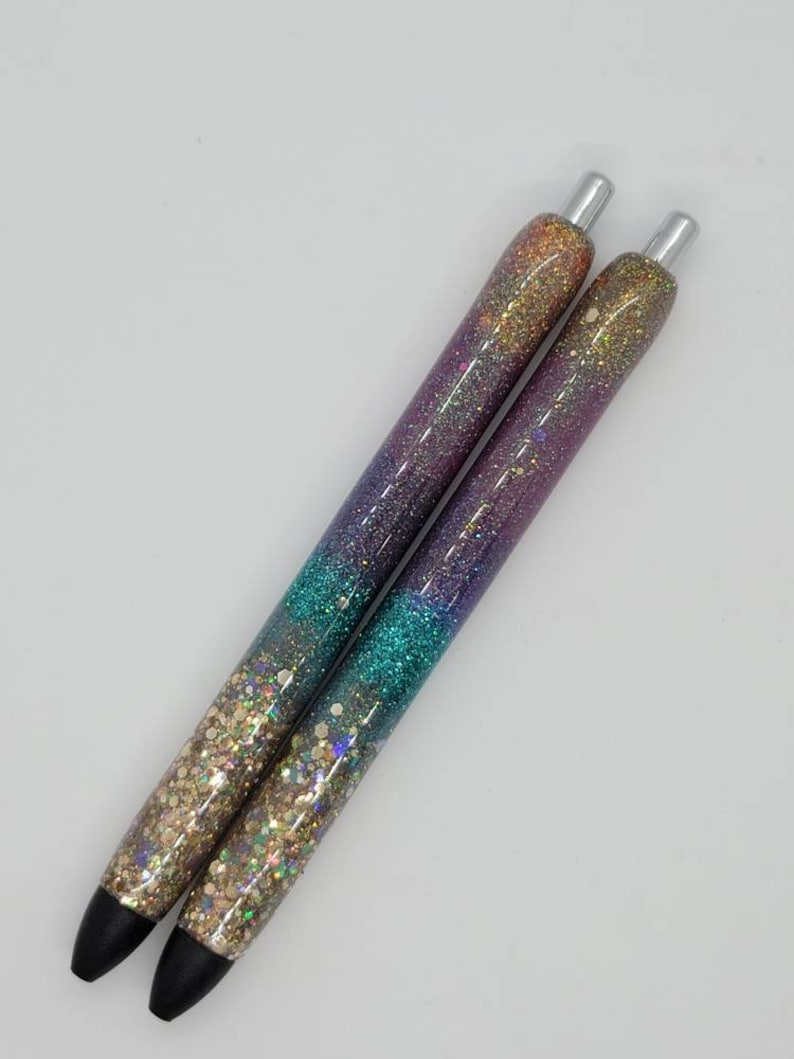 May include: Two iridescent glitter pens with a gradient of purple, teal, and gold glitter. The pens have silver caps and black ink.