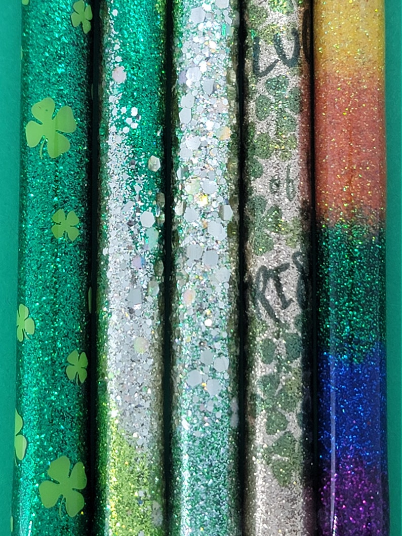 May include: Five glitter-filled tubes with different designs. The first tube is green with shamrock patterns. The second tube is silver with green glitter. The third tube is green with silver glitter. The fourth tube is silver with a shamrock pattern. The fifth tube is a gradient of orange, yellow, green, blue, and purple.