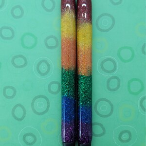 May include: Two black and silver pens with rainbow glitter. The pens are on a green background with white circles.
