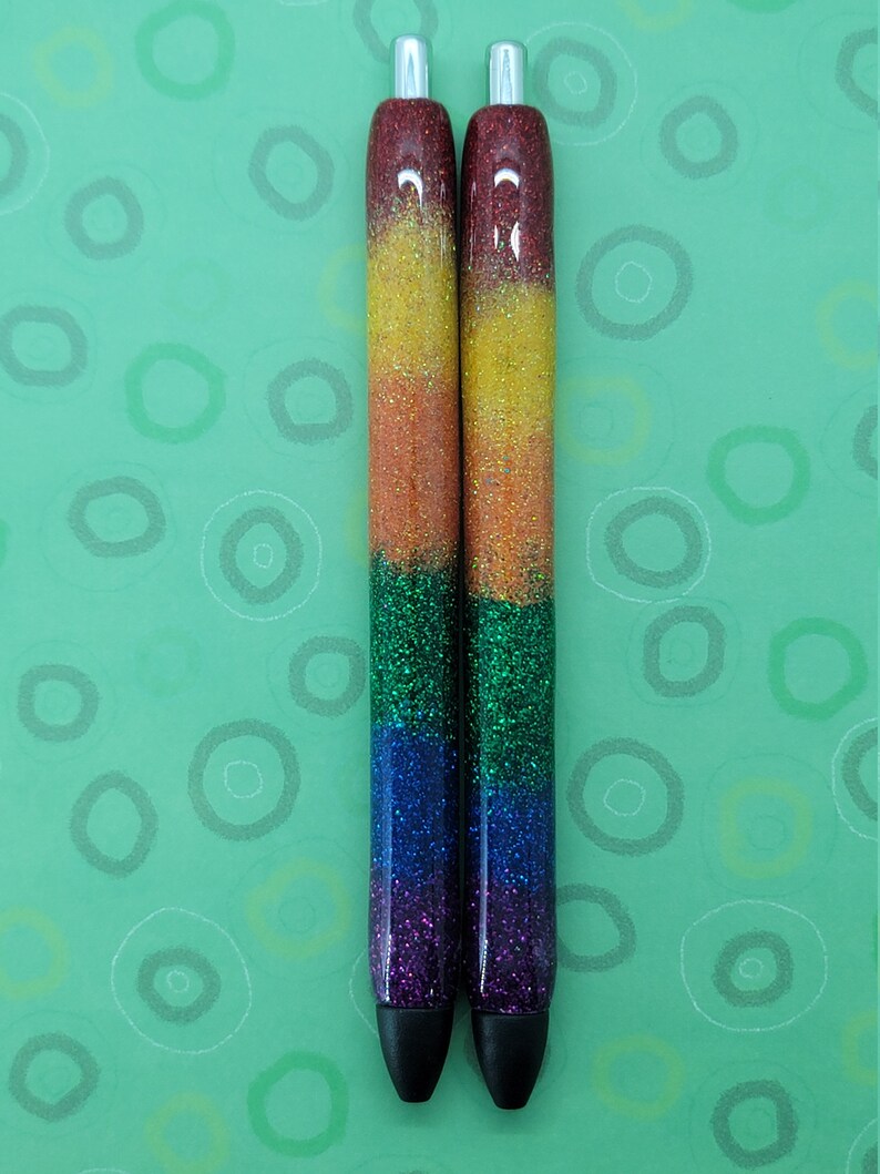 May include: Two black and silver pens with rainbow glitter. The pens are on a green background with white circles.